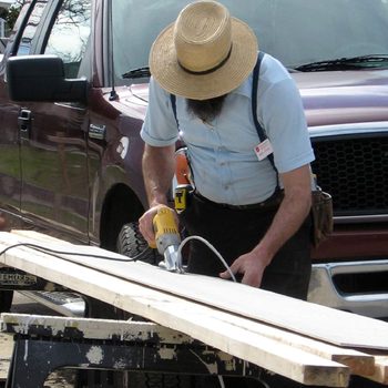 A man operates a power tool on a wooden plank, focusing on crafting. A parked truck is visible in the background.