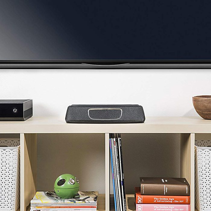 A soundbar sits on a shelf below a television, while magazines and a green decorative object are arranged nearby, creating a contemporary entertainment setup.