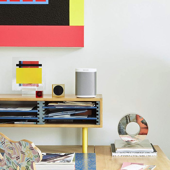 A Sonos speaker sits on a wooden table alongside colorful abstract art, decorative objects, and organized papers in a contemporary interior setting.