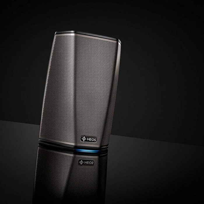 A sleek, metallic wireless speaker stands upright, reflecting light on a dark surface, emphasizing its modern design and brand logo at the base.