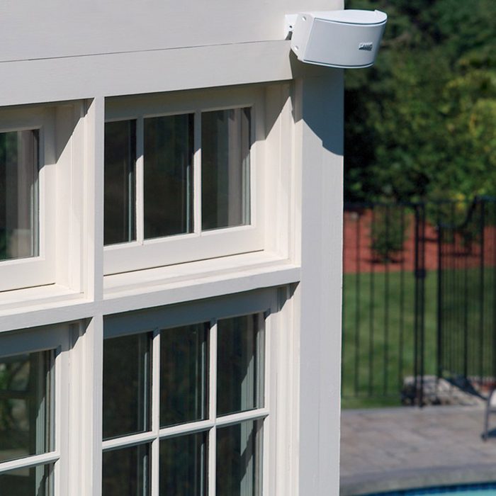 A white speaker is mounted on a light-colored building, overlooking a green lawn and a pool area surrounded by a black fence.