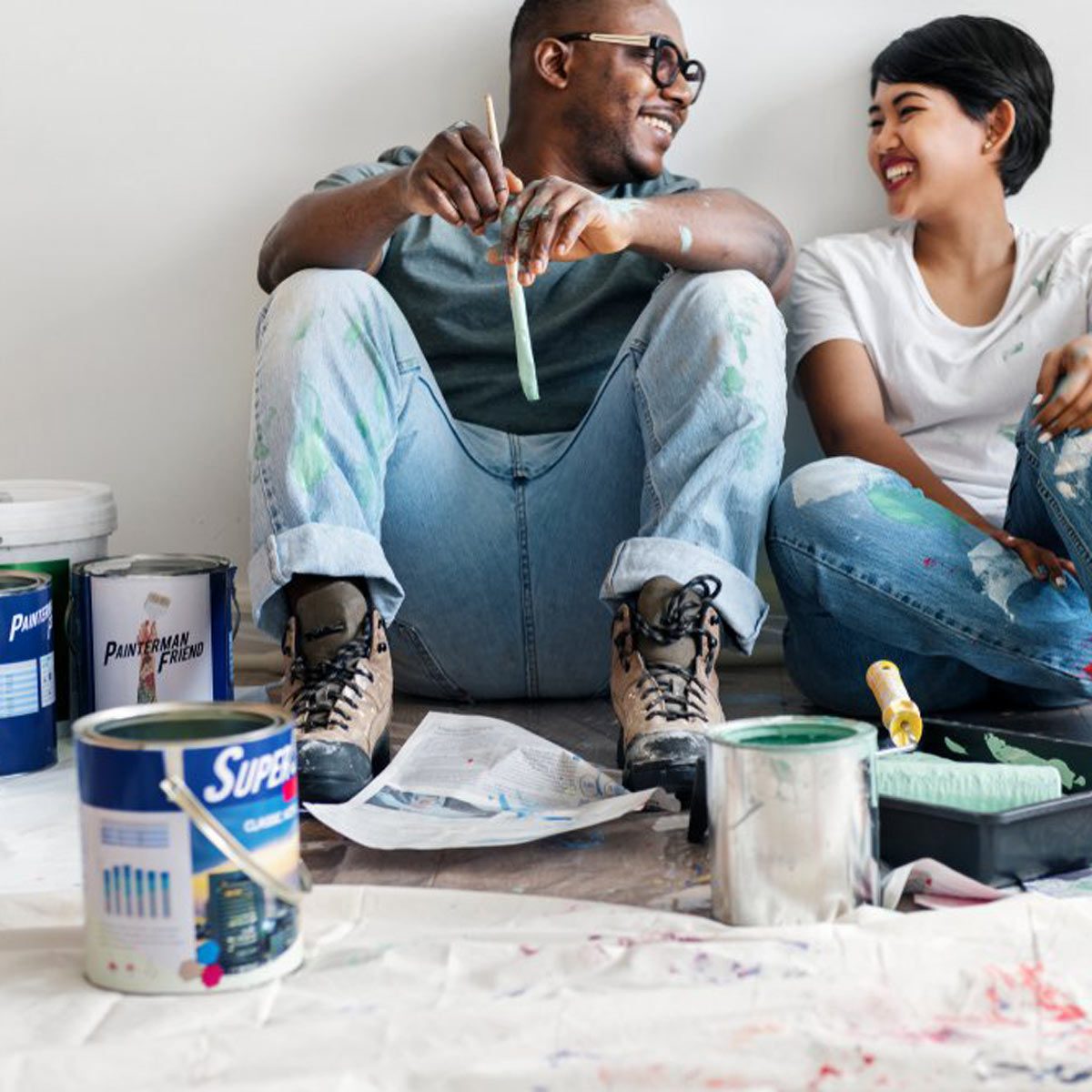 Two people sit on a floor, smiling while holding painting tools, surrounded by paint cans and a drop cloth, in a partially painted room.