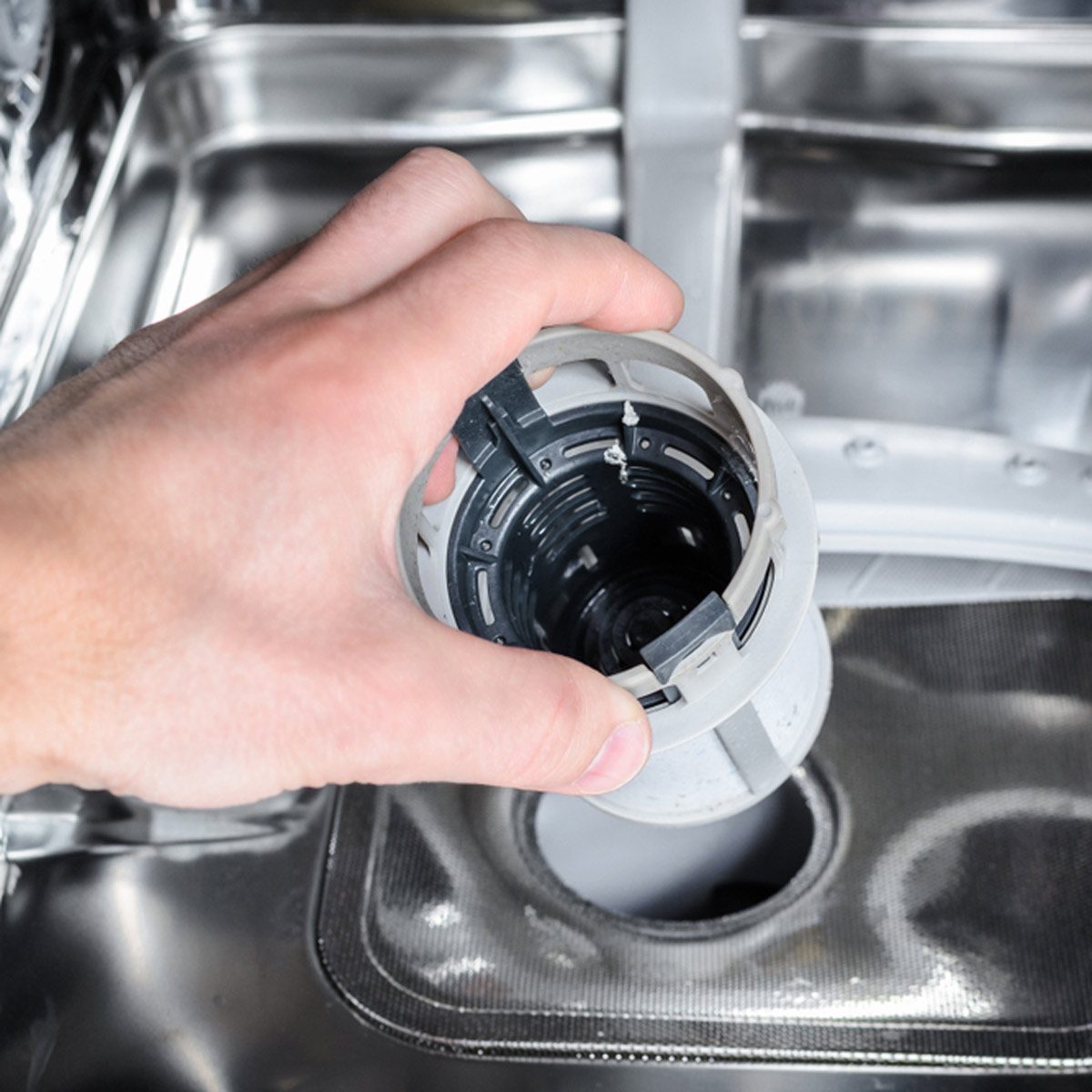 A hand holds a gray cylindrical object above a circular opening in a shiny metal surface, likely part of a kitchen appliance.