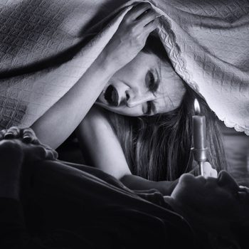 A woman screams while holding a candle underneath a blanket, looking down at a man lying below her in a dimly lit, intimate setting.