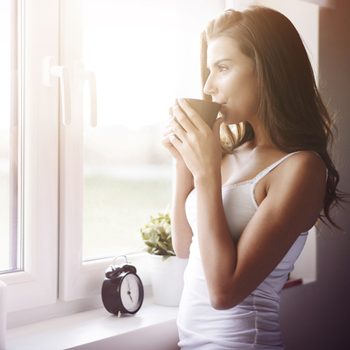A woman holds a cup, gazing thoughtfully out of a bright window, with a clock and plant nearby on a white windowsill.