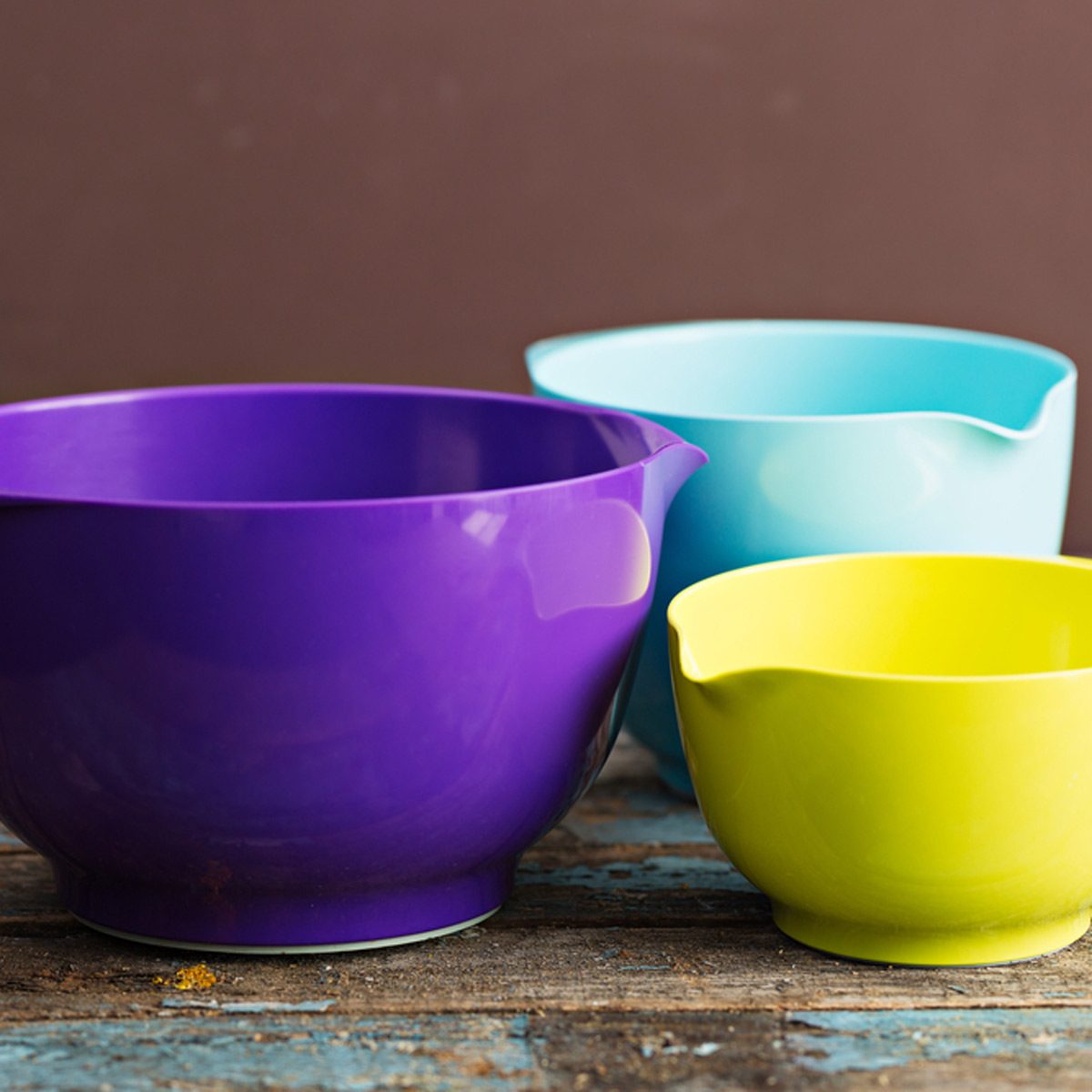 Three mixing bowls in purple, blue, and yellow rest on a rustic wooden surface, showcasing a vibrant, colorful arrangement against a neutral background.