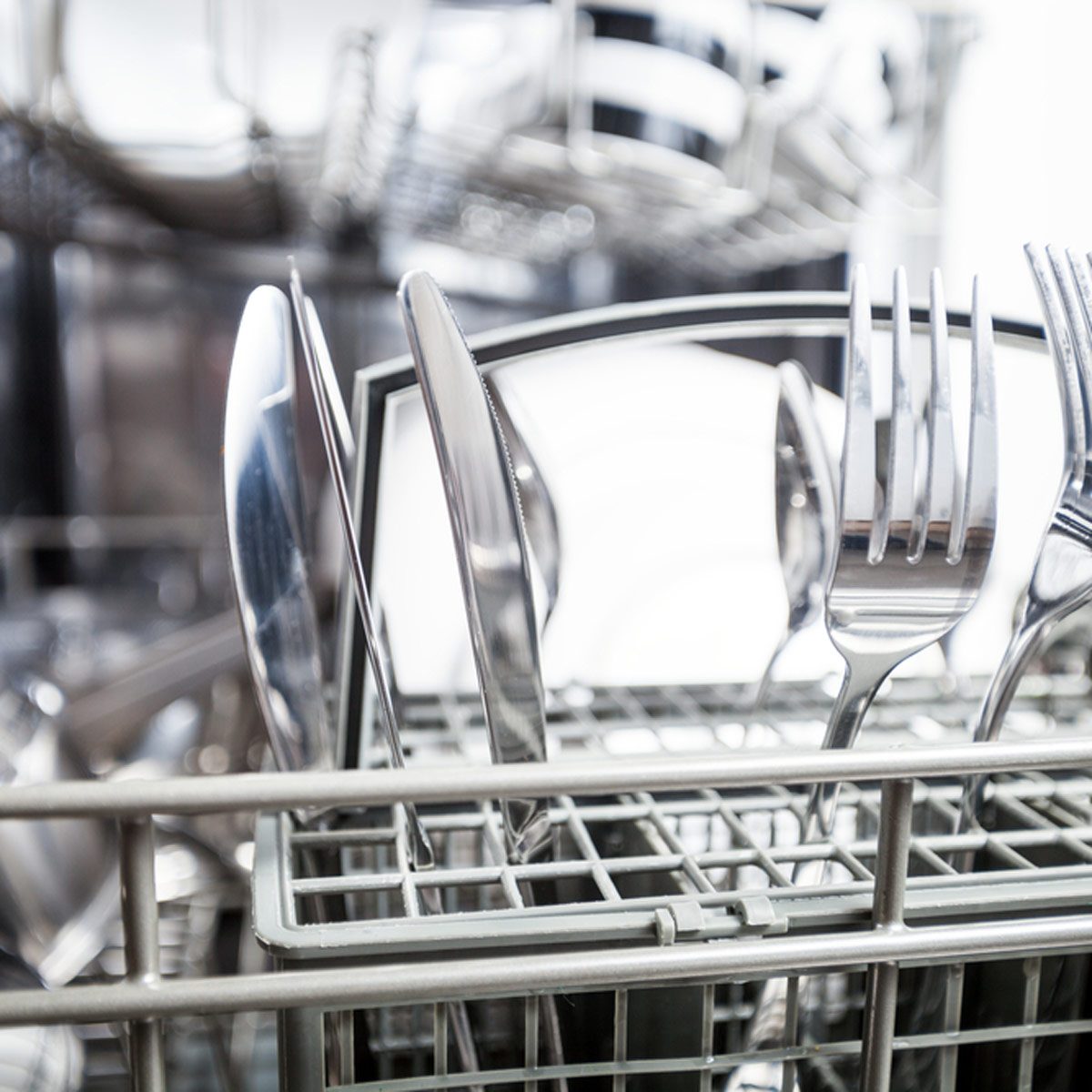 A metallic silverware basket holds various utensils, resting inside a dishwasher. The environment is clean and organized, emphasizing a kitchen appliance in use.