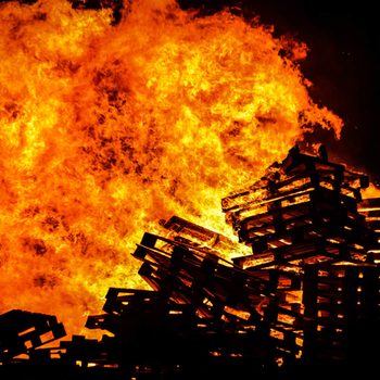 Stacks of wooden pallets are engulfed in bright orange flames, creating a vivid display against a dark background.