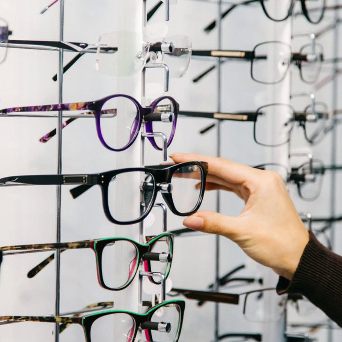 A hand reaches for a pair of black eyeglasses displayed on a rack among various styles and colors in a retail store environment.
