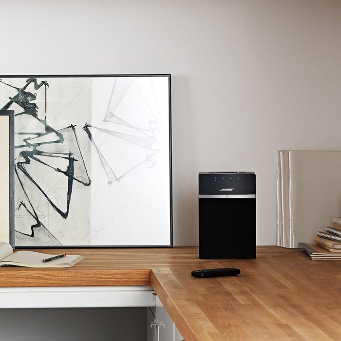A black speaker sits on a wooden desk, next to an abstract framed artwork, with an open notebook and a pencil nearby, suggesting a creative workspace.