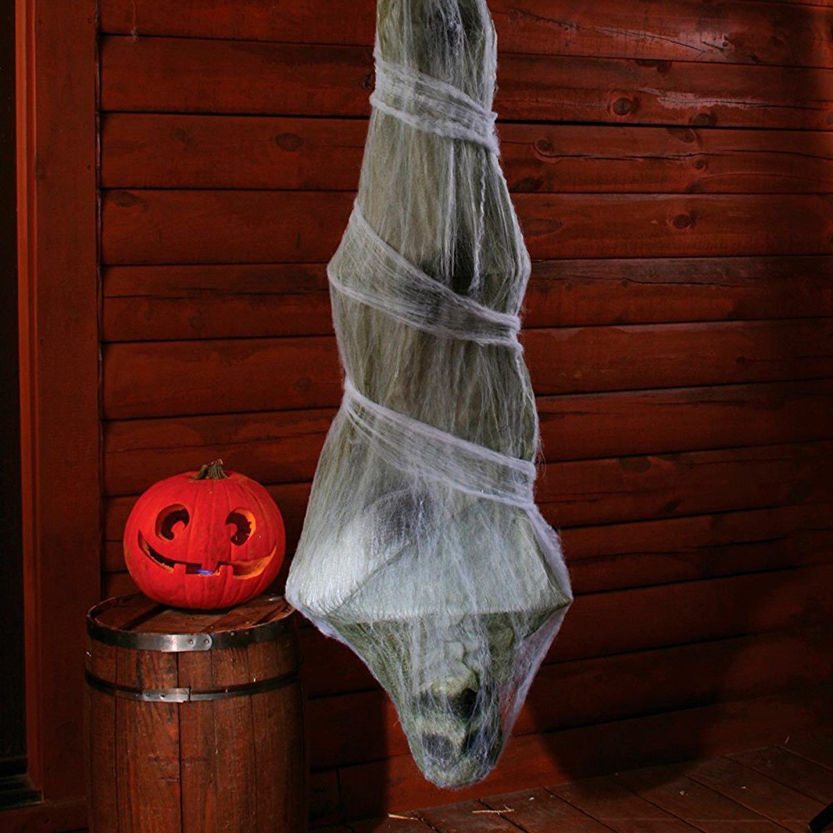 A ghostly figure is entangled in white cobwebs, suspended in a dim room alongside a carved pumpkin, creating a spooky Halloween atmosphere.