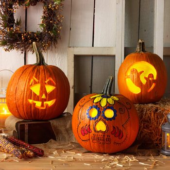 Three carved and decorated pumpkins emit light, showcasing a jack-o'-lantern, a skull design, and ghost figures, set against a rustic background with hay and decorations.