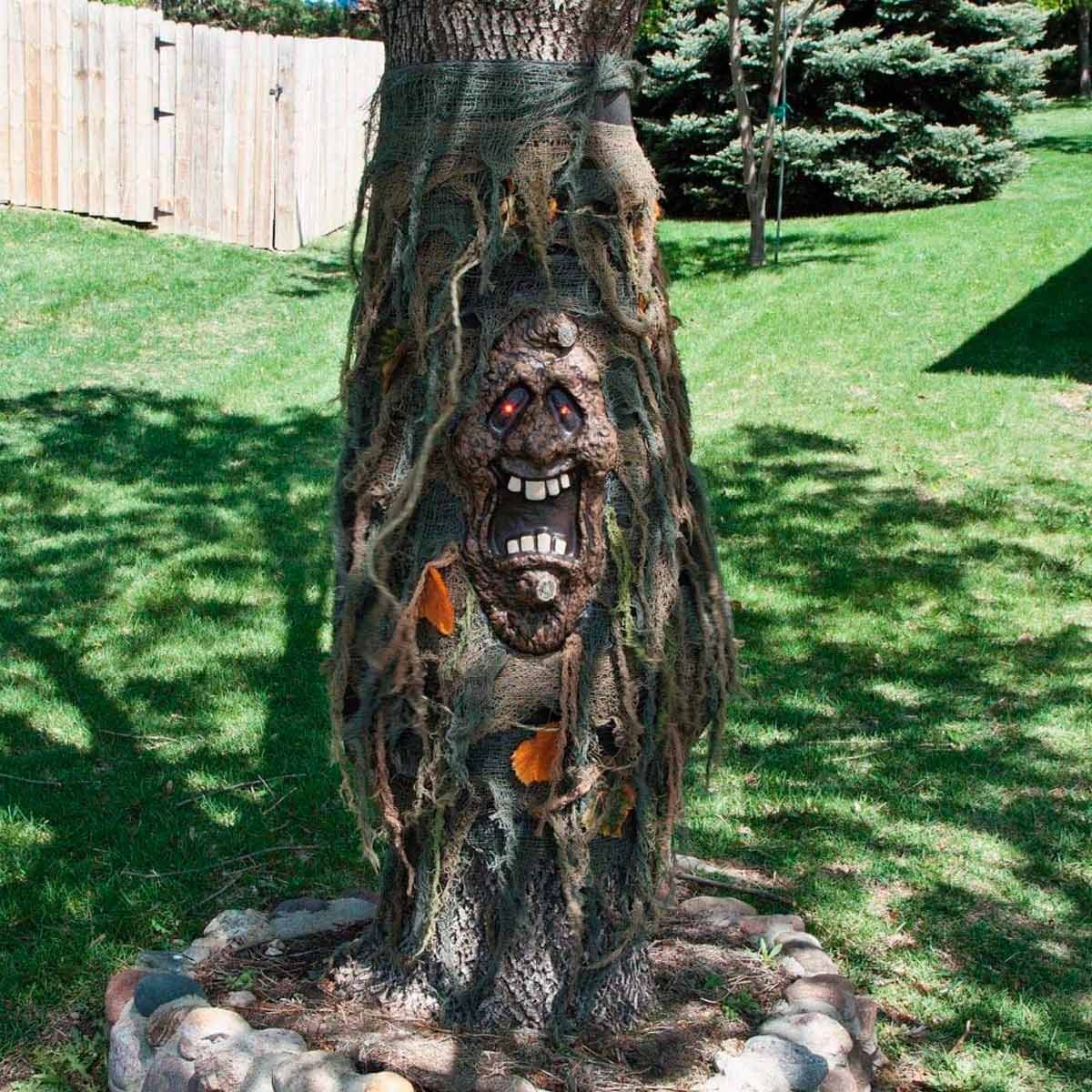 A tree trunk is partially covered in tattered fabric, revealing a carved face with exaggerated features, set against a grassy background and wooden fence.