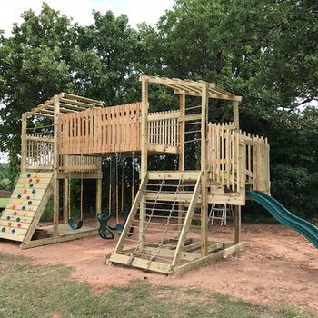 A wooden playground structure features a slide, swings, climbing wall, and various access points, set in a grassy area surrounded by trees.