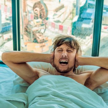 A man is lying in bed, covering his ears and grimacing, likely disturbed by loud construction noises outside a window.