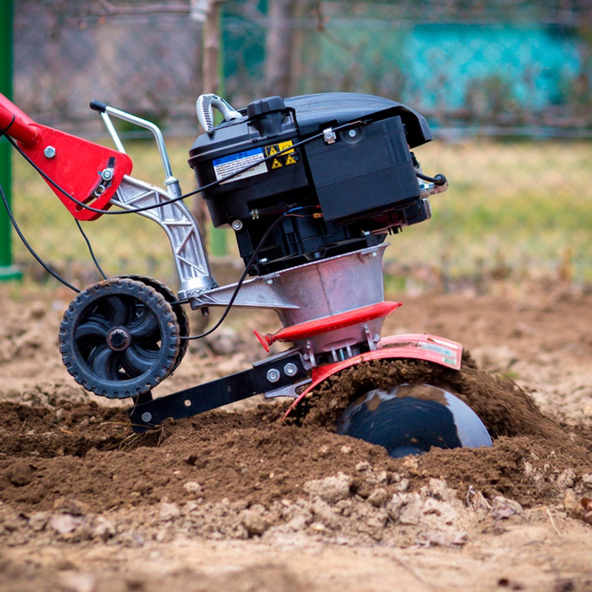 A tiller digs into the soil, turning over dirt in a garden area, with grass and a fence visible in the background.