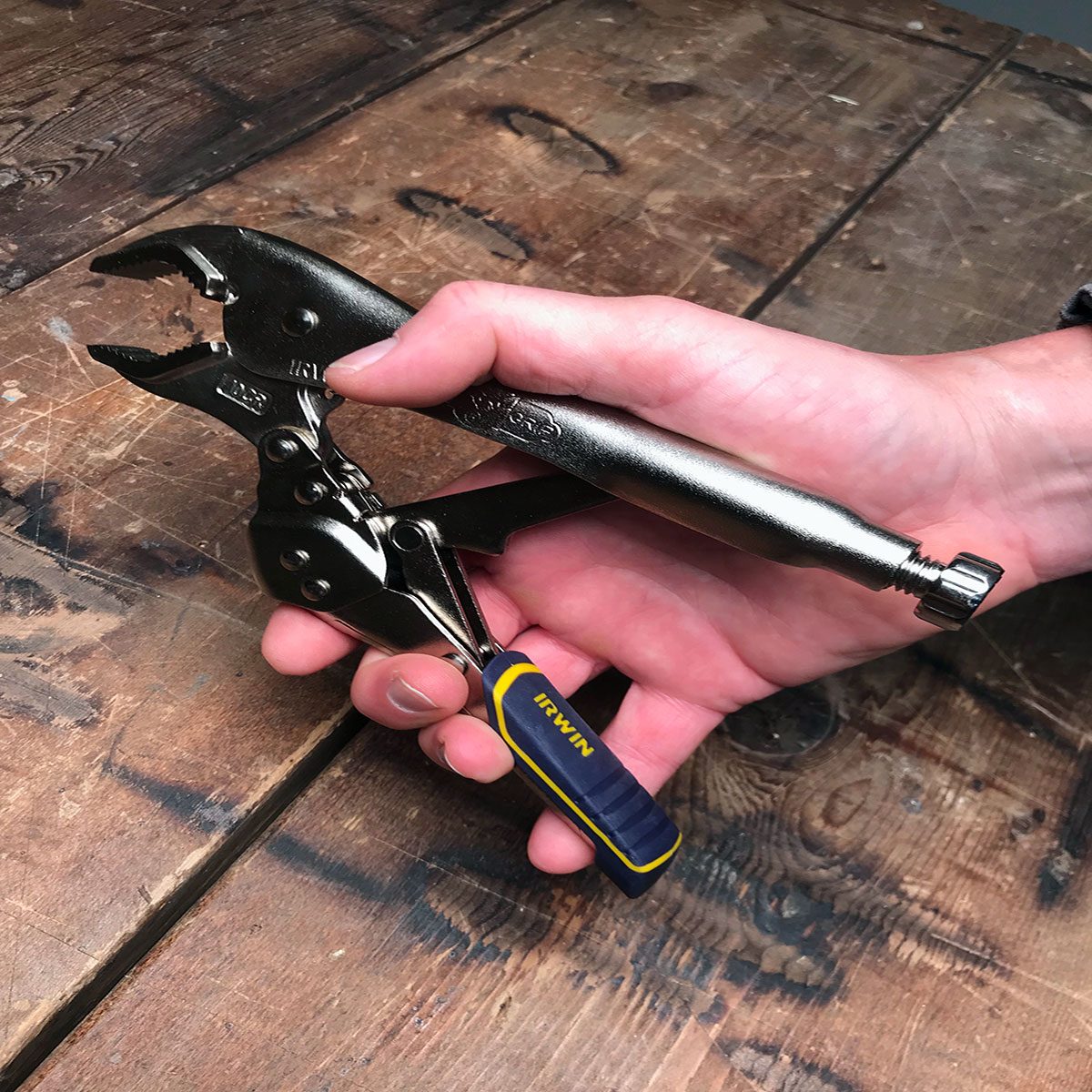 A hand grips a metallic locking plier, actively adjusting the tool's jaw against a wooden surface, showcasing a workspace setting.