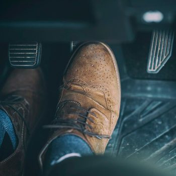 A brown dress shoe presses down on a car pedal inside a vehicle, revealing textured floor mats and surrounding controls.