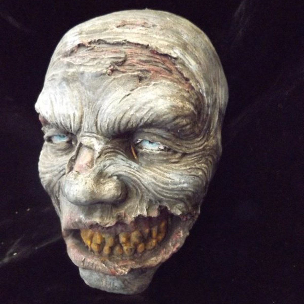 A grotesque, weathered face with deep wrinkles and jagged teeth stares unblinkingly, set against a dark background, evoking a sense of horror.