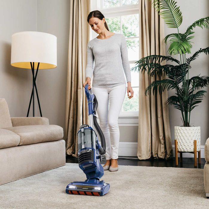 A woman vacuums a carpet with an upright vacuum cleaner in a well-lit living room, featuring a sofa, lamp, and potted plant near large windows.
