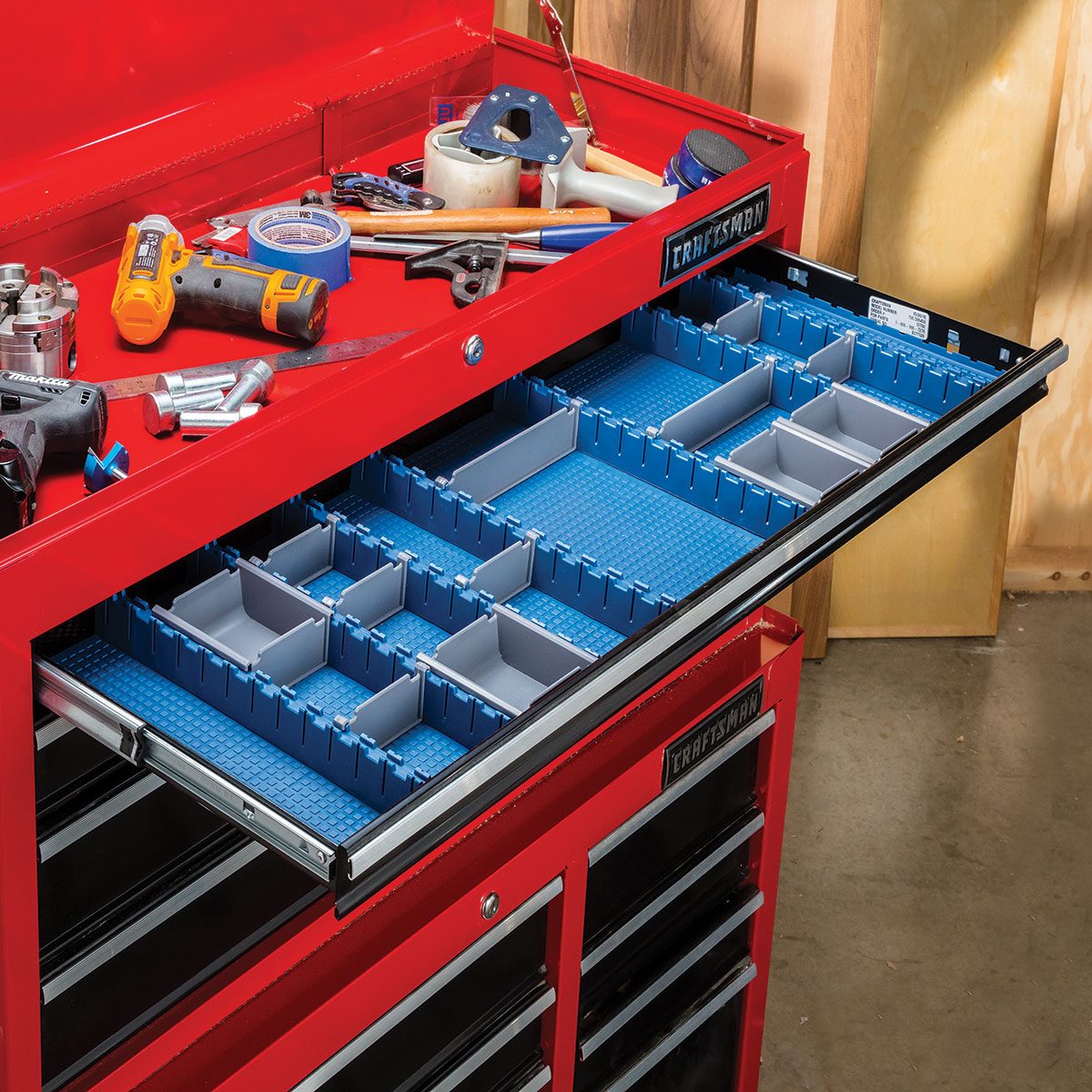 A red tool chest drawer is opened, revealing blue organizers empty of tools, surrounded by scattered tools and materials on the chest's top surface.