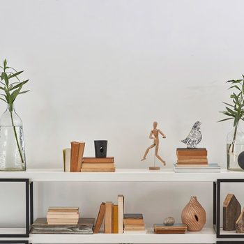 A wooden mannequin runs on a shelf, surrounded by stacked books, decorative objects, and plants in glass vases against a plain white wall.