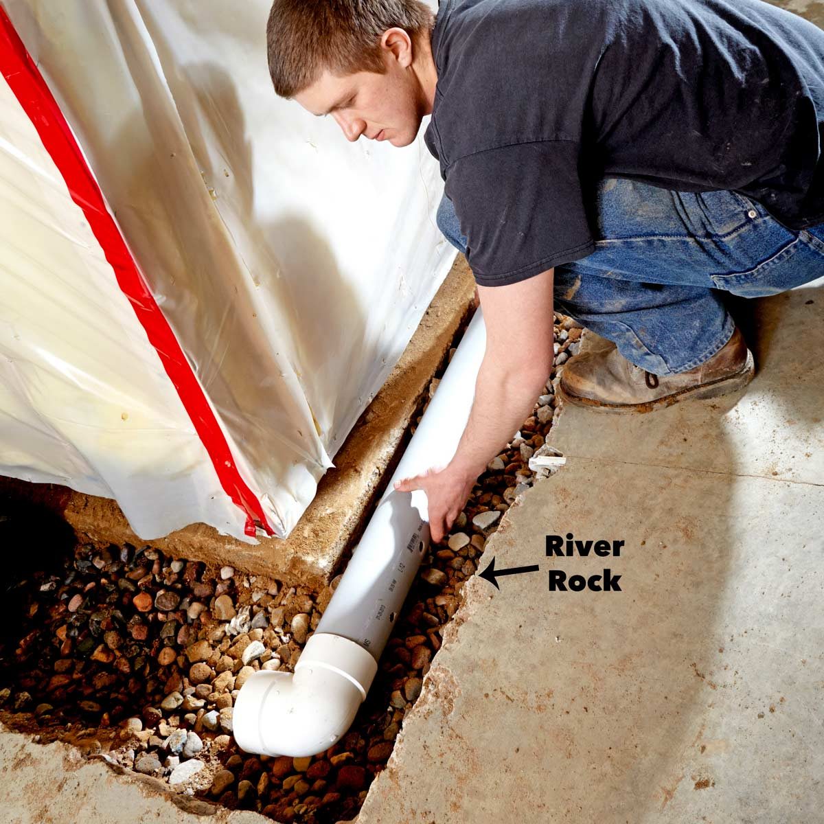 A man crouches, inspecting a white drainage pipe laid on a gravel bed beside a wall covered in plastic sheeting.