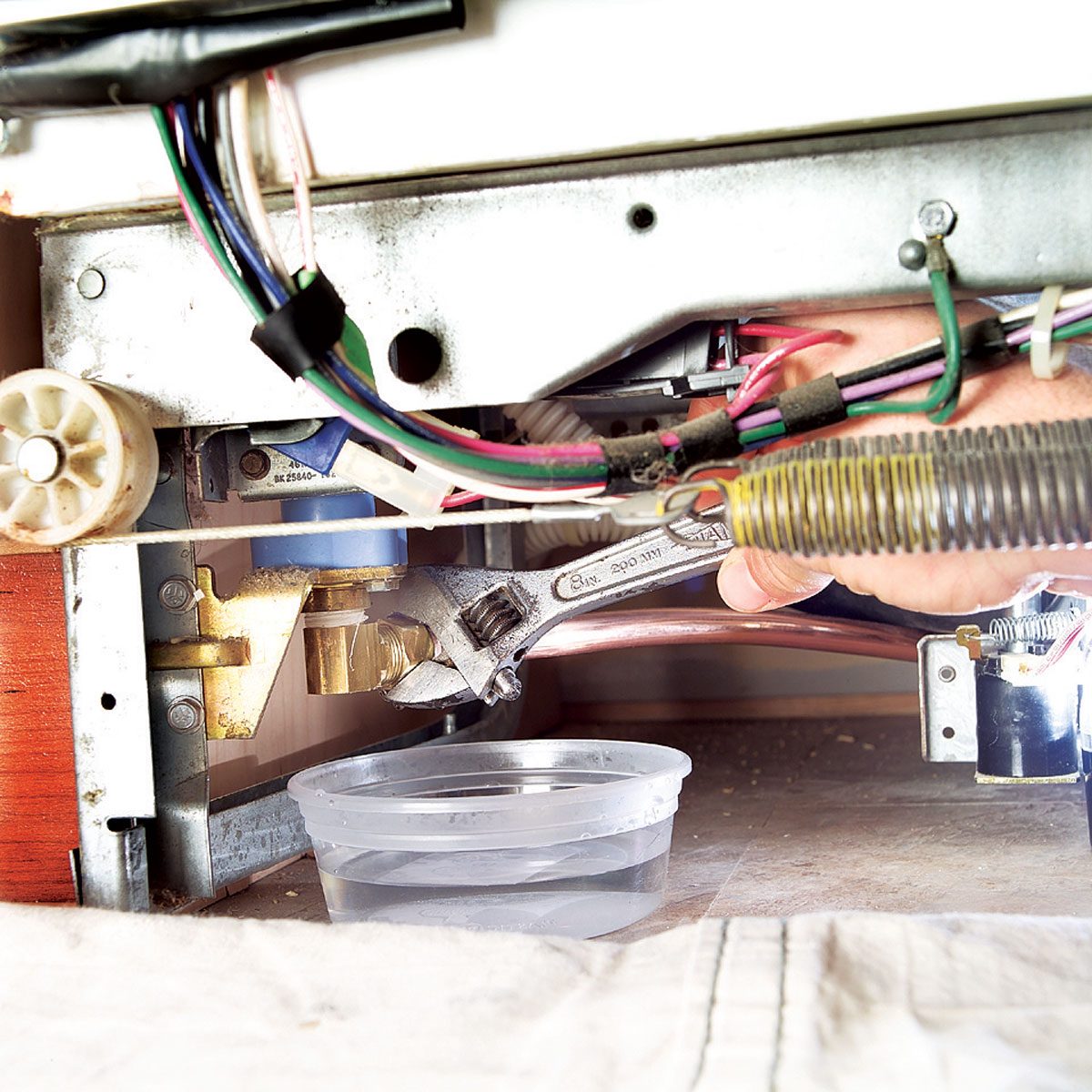 A hand holds a wrench, tightening a pipe connection under an appliance, while a cup collects water nearby in a dimly lit space.