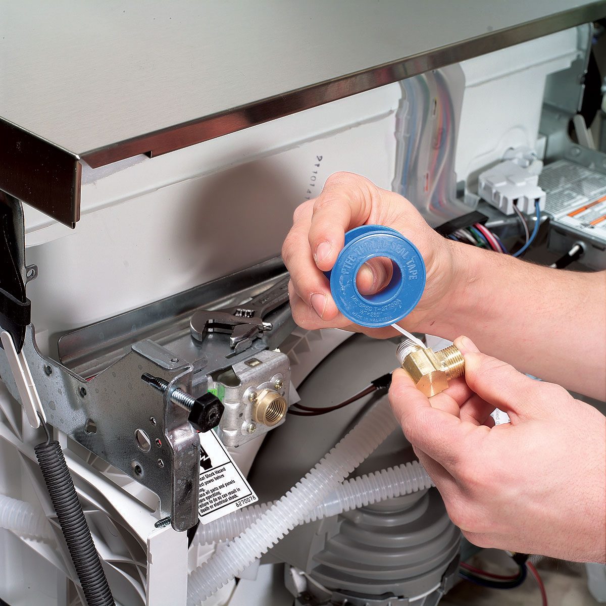 A person wraps blue tape around a brass fitting, preparing to connect it to a metal assembly in a kitchen appliance. Tools and wires are visible nearby.