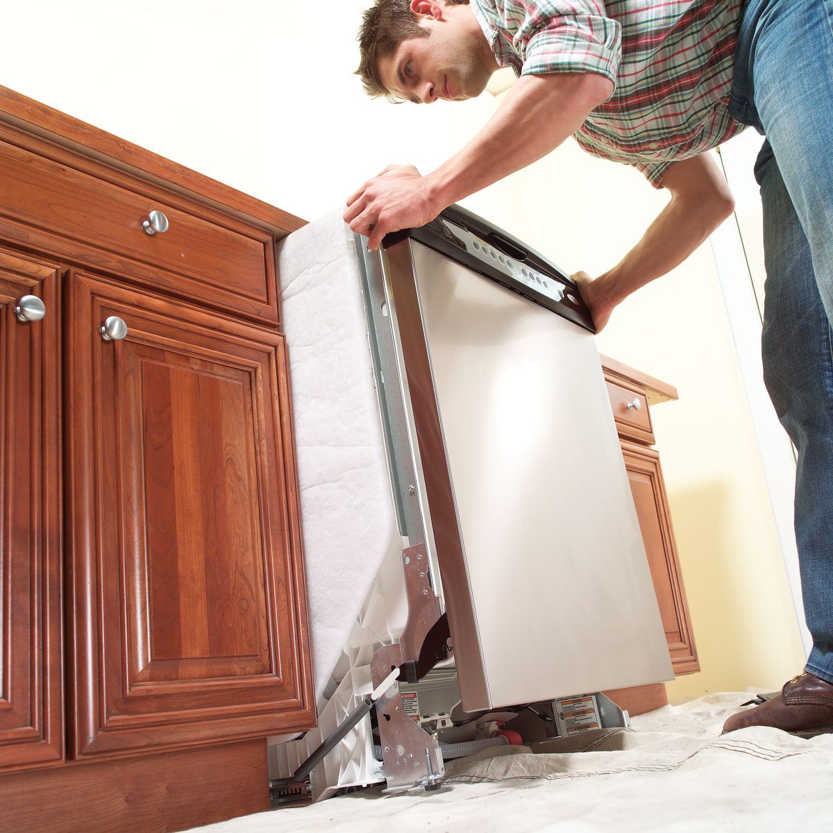 A man installs a dishwasher under wooden cabinetry, carefully aligning it while crouched on a protective sheet on the floor.