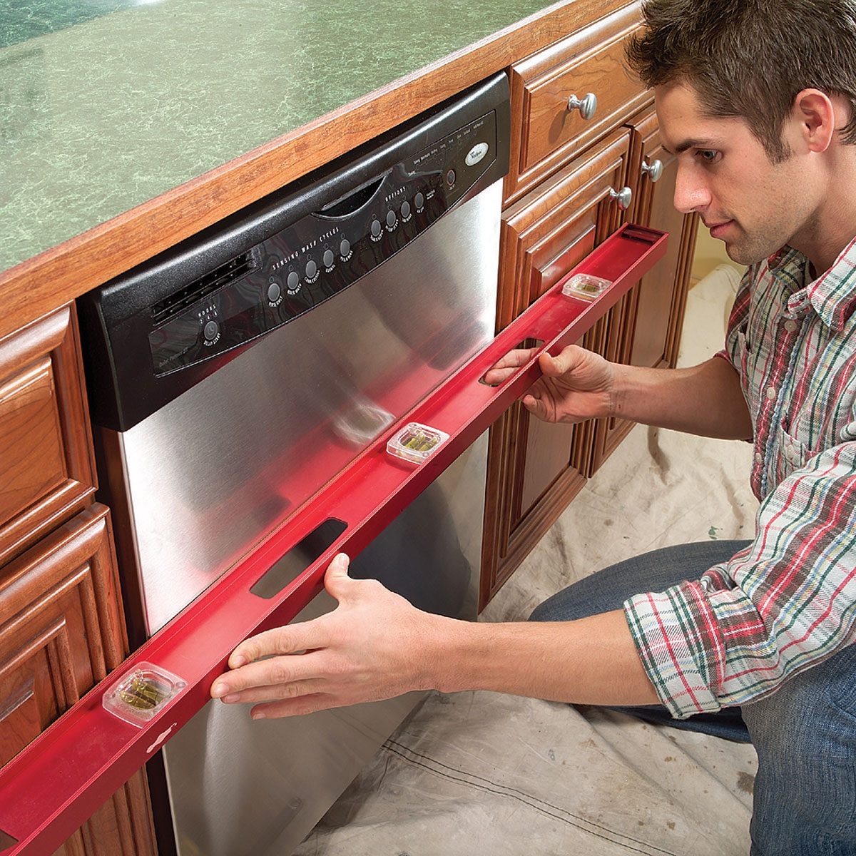 A man uses a red level tool to ensure the dishwasher is properly aligned in a kitchen with wooden cabinets and a green countertop.