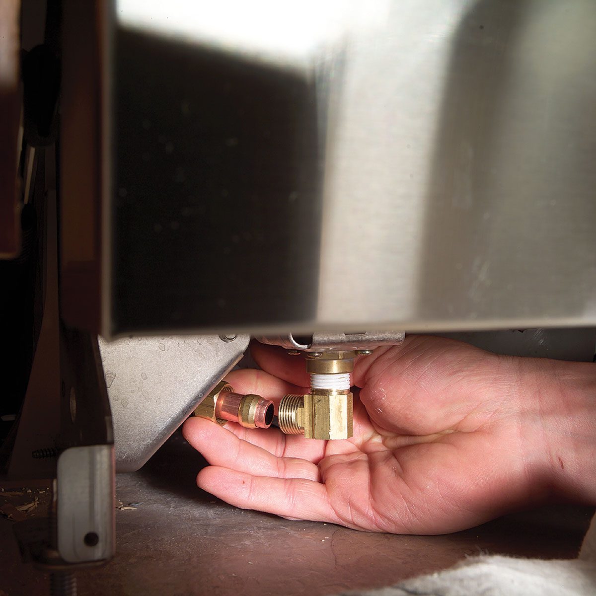 A hand is fitting a brass connector under a machine, surrounded by metal surfaces and a dimly lit, cluttered work area.