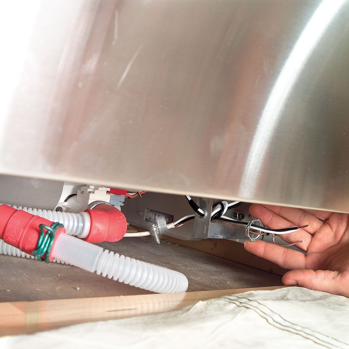 A hand reaches toward plumbing and electrical components beneath a cabinet, with red hoses and wires visible on a wooden surface.