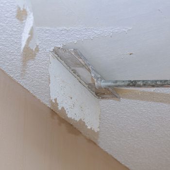 A paint scraper removes texture from a ceiling, with flakes of material falling. The background shows a flat, untextured wall in a light color.