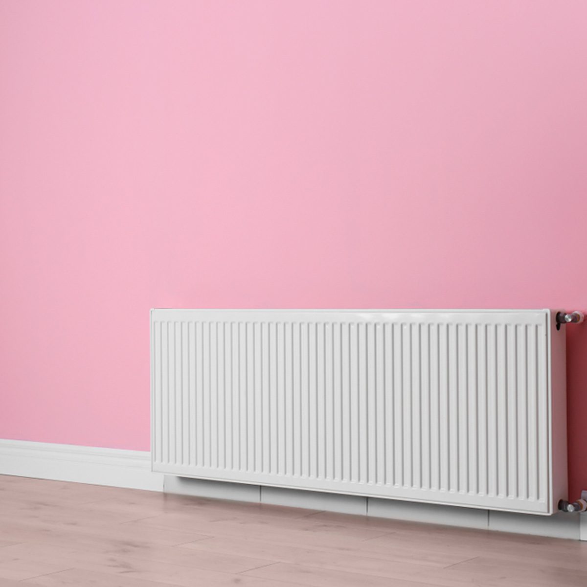 A white radiator runs along a smooth floor, warming a room with a soft pink wall, creating a bright and cozy atmosphere.
