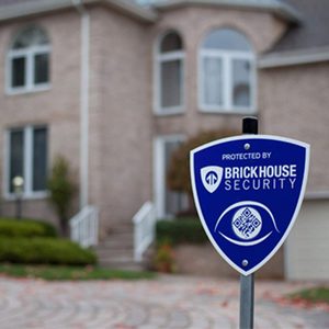 A security sign stands in front of a brick house, indicating protection by Brickhouse Security, with a notable QR code displayed on the sign.
