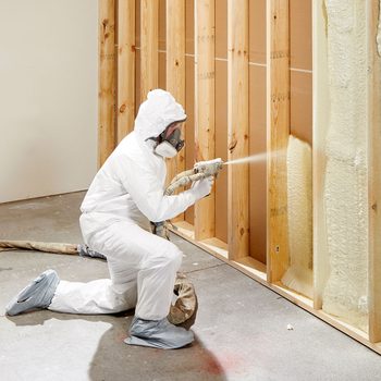 A person in protective gear sprays insulation onto wooden wall framing while kneeling on a concrete floor in an unfinished indoor space.