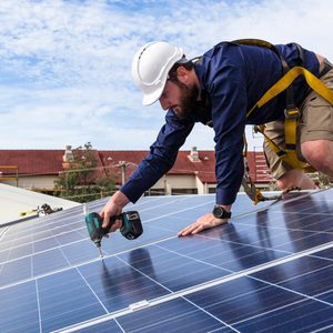 A man in a hard hat uses a power drill to secure solar panels on a rooftop, with residential buildings and trees visible in the background.