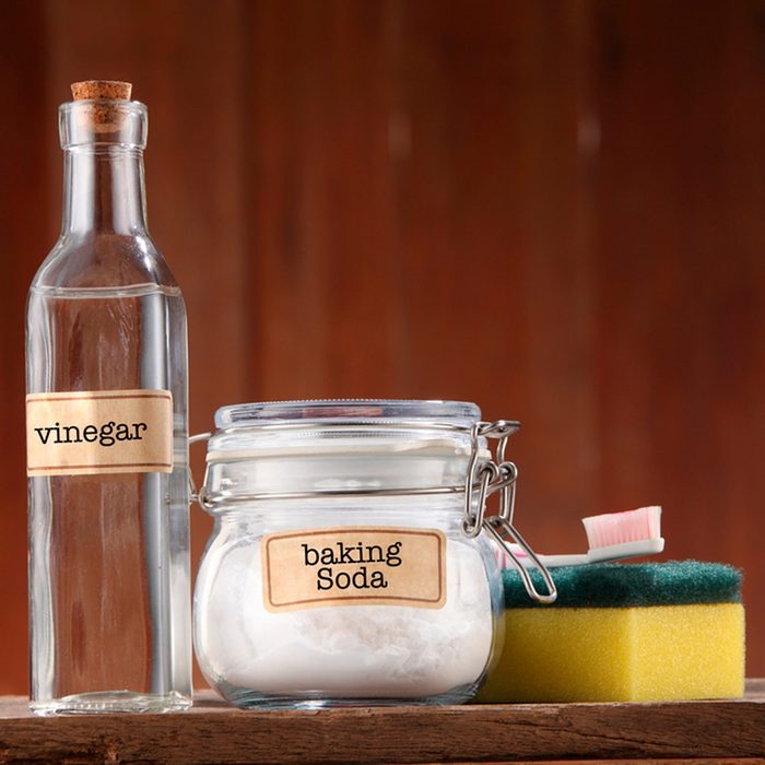 A glass bottle of vinegar and a jar of baking soda sit on a wooden surface, accompanied by a yellow sponge and a toothbrush.