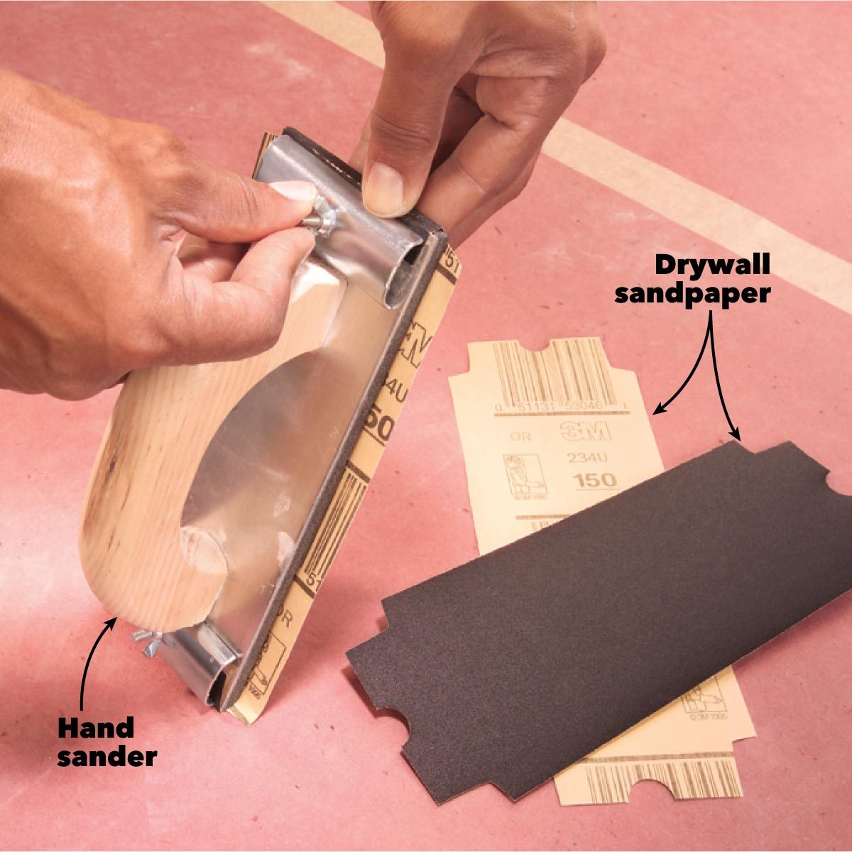 A hand sander holds a piece of drywall sandpaper, preparing to attach it on a flat, reddish surface, with packaging visible nearby.
