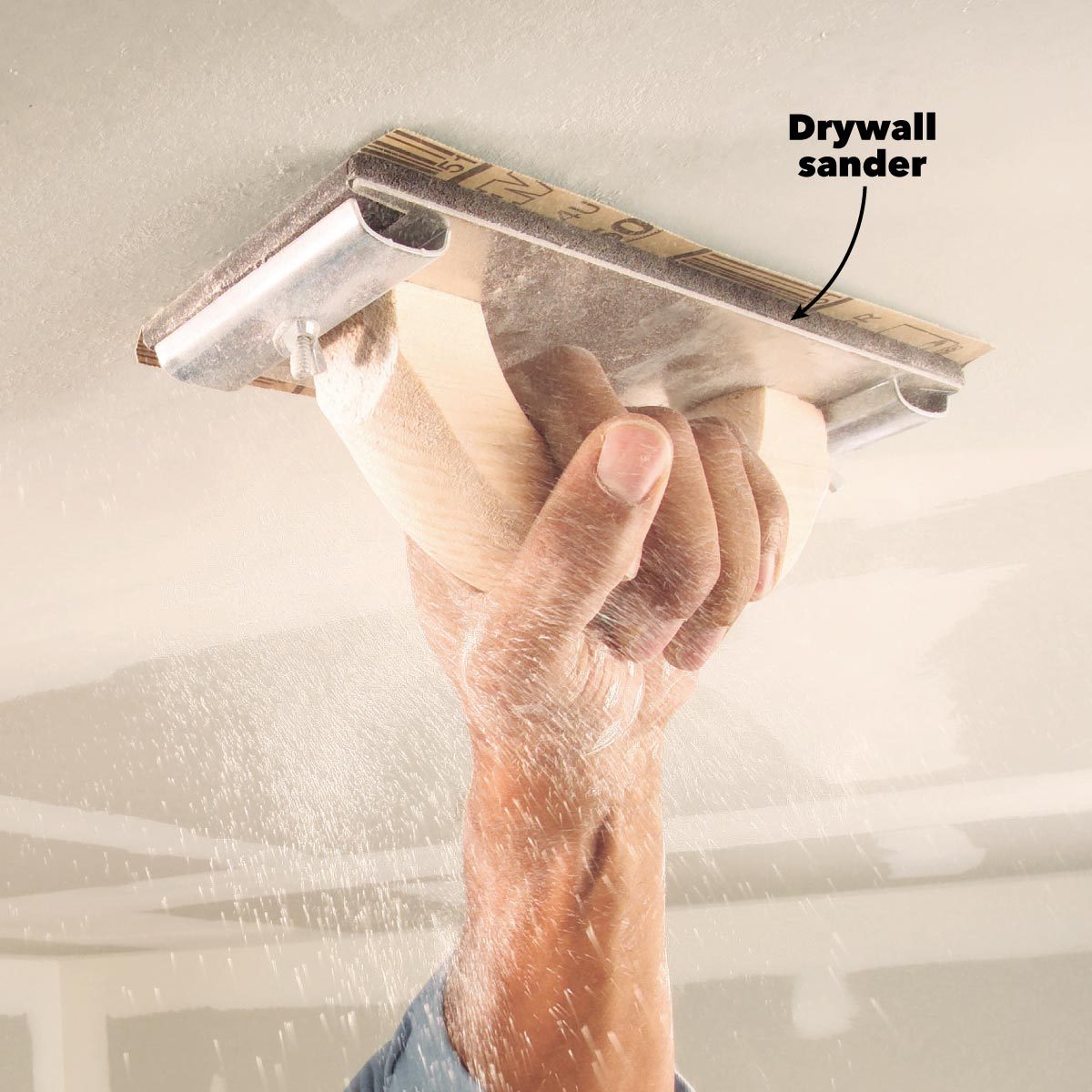 A hand holds a drywall sander, smoothing a ceiling surface while fine dust particles disperse in the air.