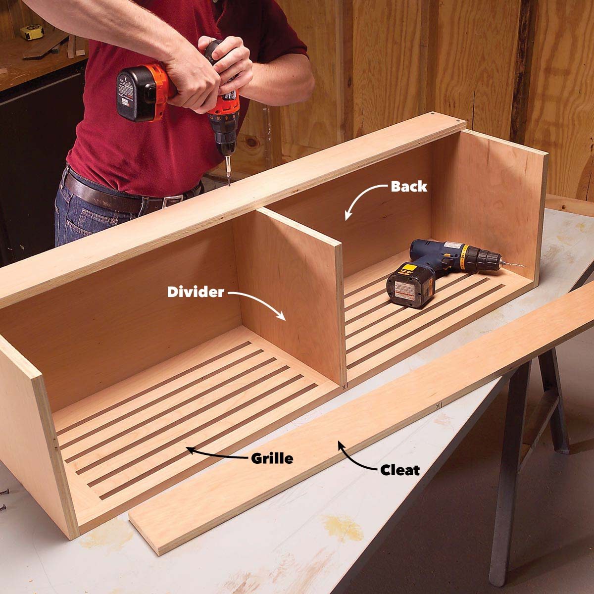 A person uses a drill to assemble a wooden structure with a divider and slatted bottom, surrounded by tools and parts on a workbench.
