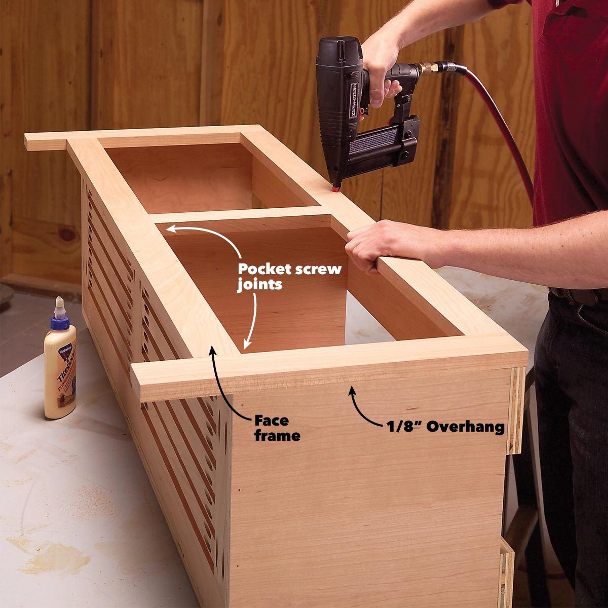 A person uses a nail gun to assemble a wooden frame with pocket screw joints on a work surface, surrounded by wooden walls in a workshop.