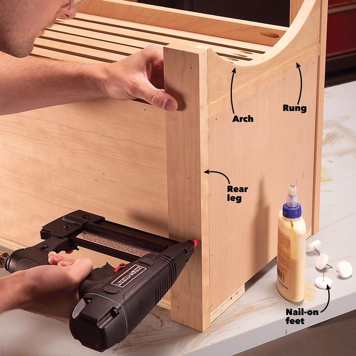 A person uses a nail gun to attach a wooden piece to a project, surrounded by glue and nail-on feet components on a work surface.