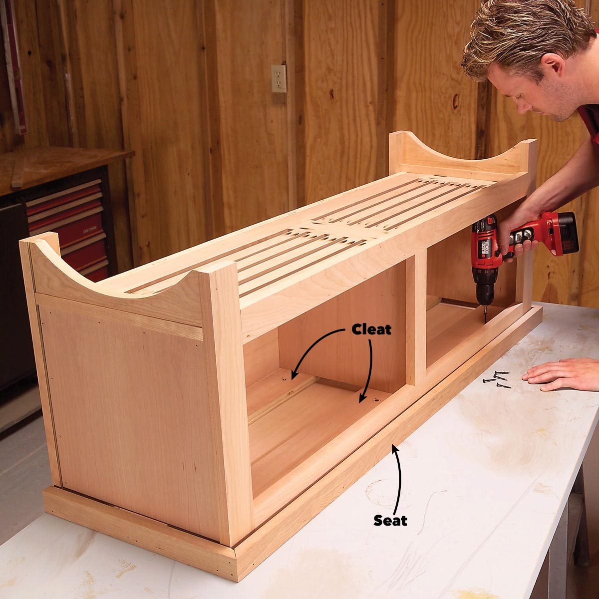 A wooden bench is being assembled by a person using a cordless drill in a workshop, featuring slats on top and storage compartments below.