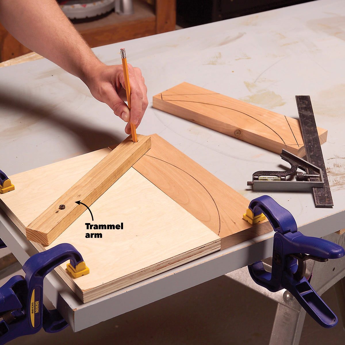 A hand holds a pencil while marking a curve on wood using a trammel arm, positioned on a workbench with other tools nearby.