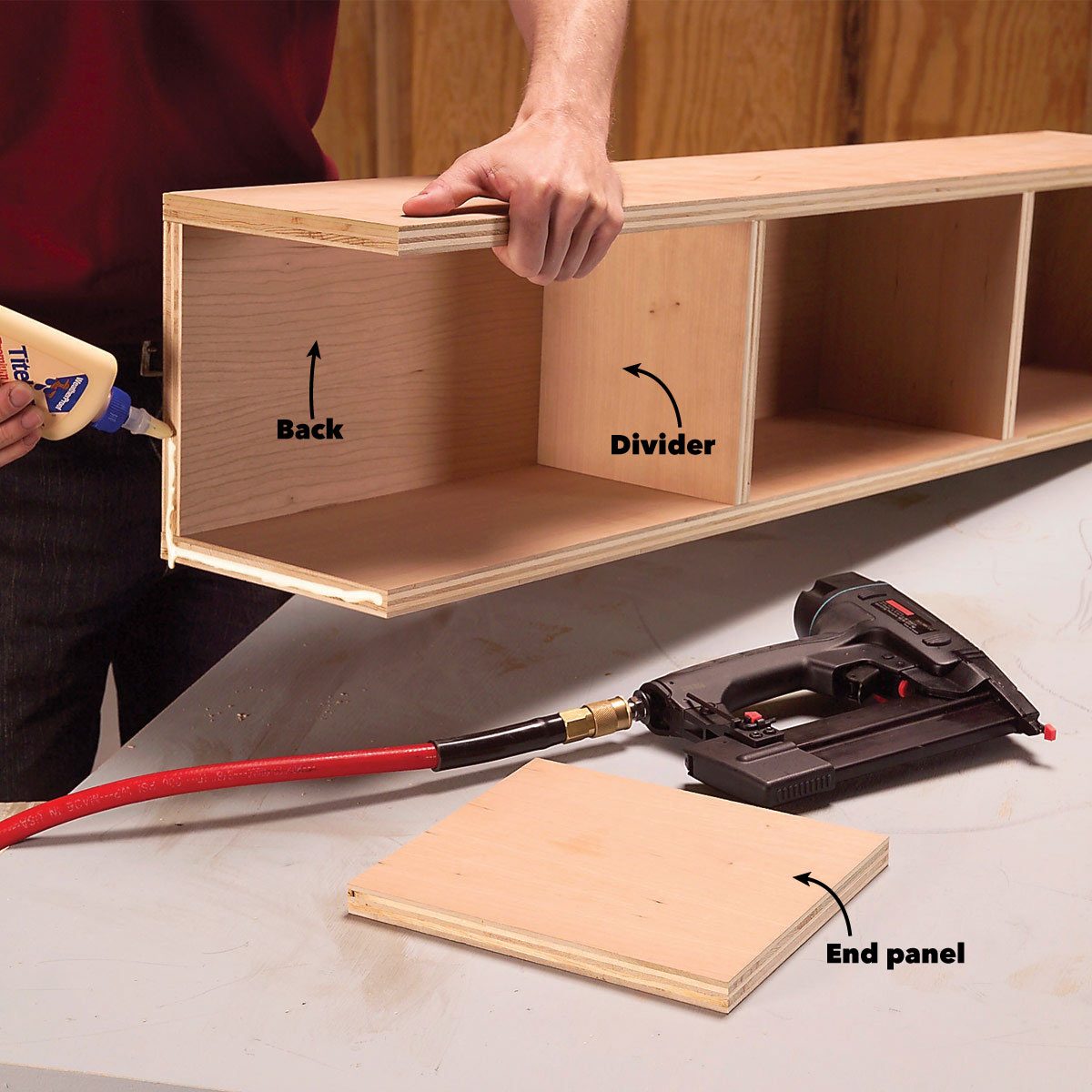 A person applies glue inside a wooden shelf frame, preparing to insert a divider, with an end panel and a nail gun on a work surface.