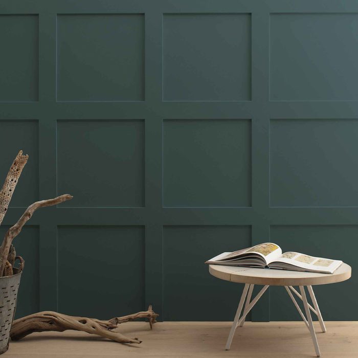 A round table holds an open book, while driftwood and a basket rest against a dark green paneled wall in a minimalist interior.