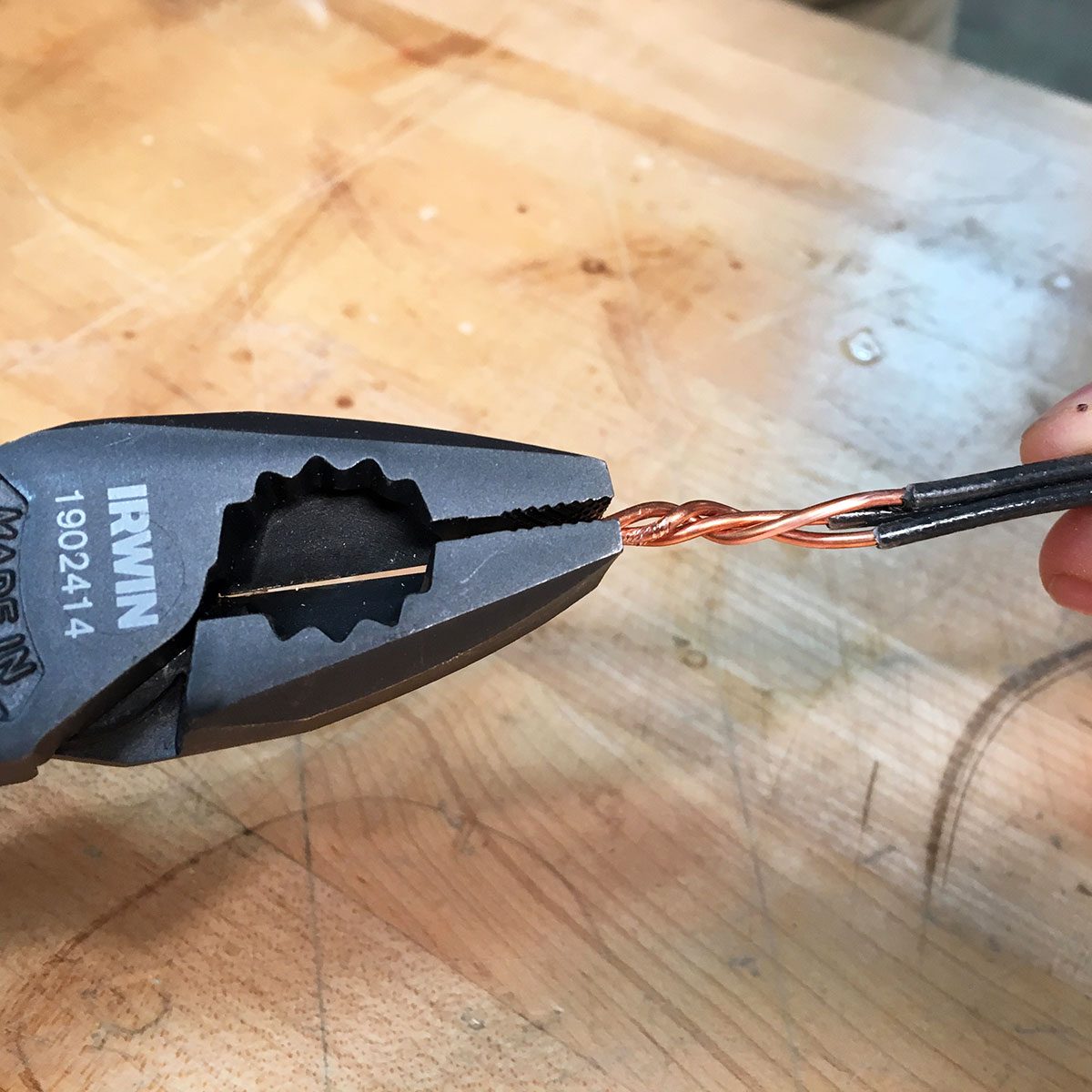 Twisting wires with a linesman's pliers. A pair of pliers grips twisted copper wires, preparing for manipulation on a wooden work surface.