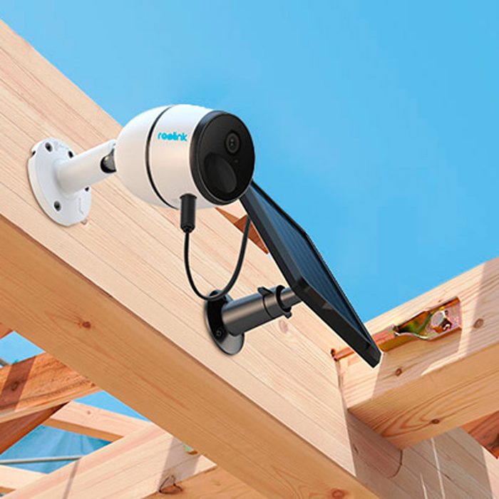 A surveillance camera is mounted on wooden beams, capturing the surroundings while connected to a solar panel for power under a clear blue sky.